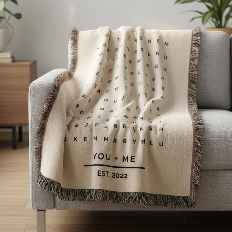 May include: Cream-colored throw blanket with a black letter design and fringe trim. The blanket features the text "YOU + ME" and "EST. 2022". The blanket is draped over a gray sofa.