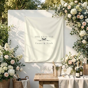 Könnte beinhalten: Ein cremefarbenes Banner mit dem Text „SHE SAID YES“ und den Namen „Emma & Noah“ wird angezeigt. Eine Schleifengrafik ist enthalten. Die Szene ist für eine Hochzeit vorbereitet, mit Champagner, Gläsern und Blumengestecken.