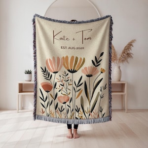 Könnte beinhalten: Beige gewebte Decke mit einem Blumendesign in Pfirsich-, Orange- und Grüntönen. Die Decke hat einen Fransenrand und enthält die Namen "Kate + Tom" und das Datum "EST. AUG 2020".