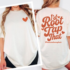 Puede incluir: Camiseta color crema con un coraz&oacute;n color &oacute;xido en la parte delantera. La parte trasera de la camiseta presenta el texto "I'd Root Tap That" en una fuente estilizada color &oacute;xido, con el identificador de Instagram @katemahoneyhair debajo.