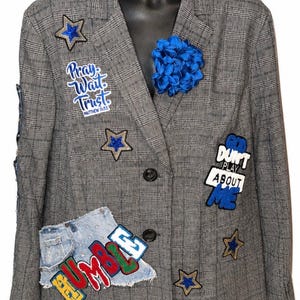 May include: A grey plaid blazer featuring various patches including blue stars, a blue fabric flower, and text patches reading Pray Wait Trust, GOD DONT PLAY ABOUT ME, and HUMBLE, along with a denim fabric patch on the lower left side.