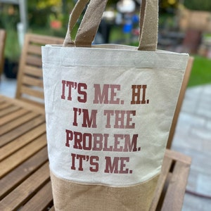 Könnte beinhalten: Eine weiße Canvas-Tragetasche mit braunen Henkeln und einem braunen Juteboden. Die Tasche hat den Text "It's me. Hi. I'm the problem. It's me." in Rot auf der Vorderseite aufgedruckt.