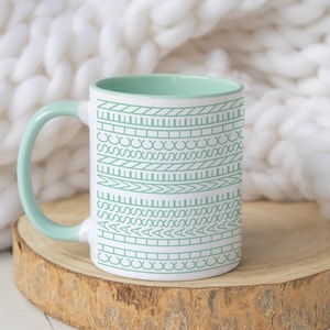 Könnte beinhalten: Weißer Keramikbecher mit mintgrünem Henkel und Innenraum. Der Becher hat ein sich wiederholendes Muster aus geometrischen Formen in Mintgrün. Der Becher steht auf einer Holzscheibe, im Hintergrund eine weiße, grob gestrickte Decke.