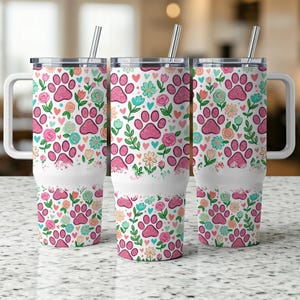May include: Three white travel mugs with handles and lids. The mugs feature a repeating pattern of pink paw prints, flowers, and hearts on a white background. Each mug includes a silver straw. The mugs are designed for cold beverages.