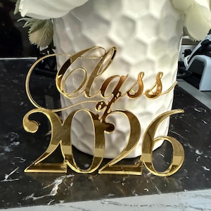 以下が含まれることがあります： 「Class of 2026」の文字が筆記体で書かれた、金色の反射性アクリルケーキトッパー。白い花が入った白い花瓶の前に置かれています。