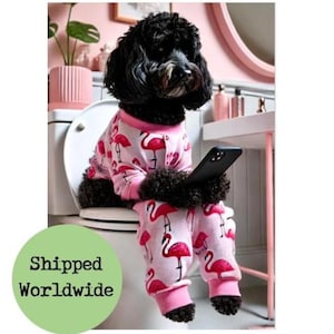 May include: A black dog wearing pink flamingo pajamas, sitting on a toilet and holding a black smartphone. The pajamas have a pink background with red flamingos. The dog is in a pink bathroom setting.