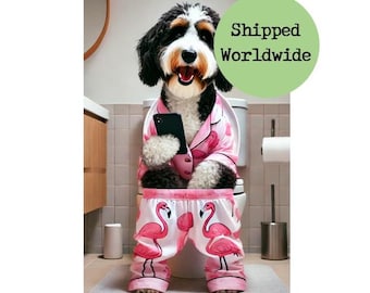 Op de afbeelding: Een zwart-witte hond in een roze pyjama met flamingo print zit op een witte toiletbril. De hond houdt een zwarte smartphone in zijn poten. De hond is in een badkamer met een witte betegelde vloer en een houten kast.