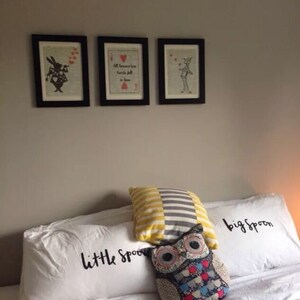 Pu&ograve; includere: Due federe bianche con il testo "little spoon" e "big spoon" su di esse. Un cuscino a righe giallo e grigio e un cuscino a forma di gufo colorato sono sopra le federe. Tre stampe in bianco e nero incorniciate sono sul muro dietro i cuscini.