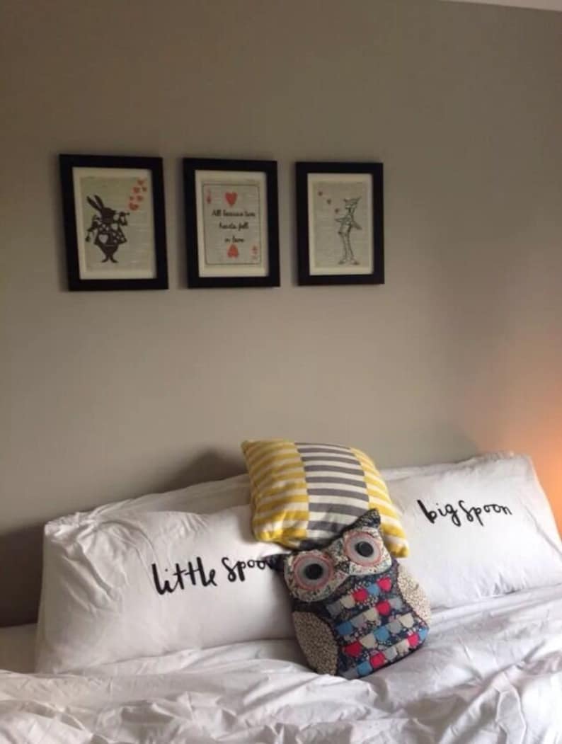 Pu&ograve; includere: Due federe bianche con il testo "little spoon" e "big spoon" su di esse. Un cuscino a righe giallo e grigio e un cuscino a forma di gufo colorato sono sopra le federe. Tre stampe in bianco e nero incorniciate sono sul muro dietro i cuscini.