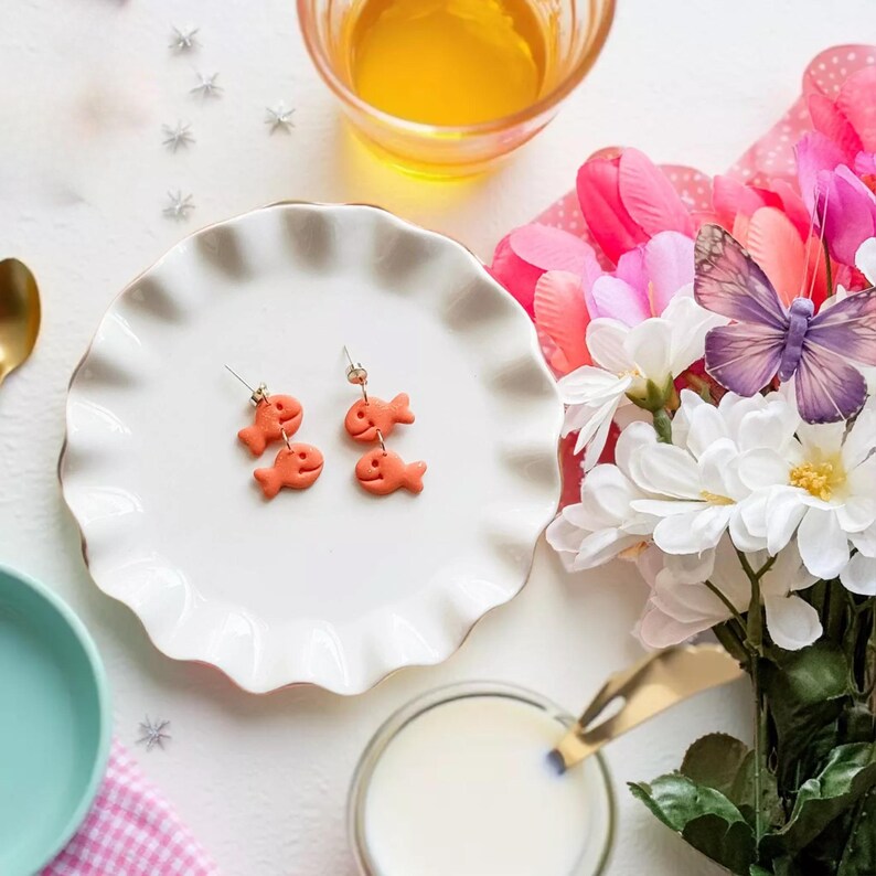 a white plate with two orange fish-shaped earrings, a glass of milk, a teapot, a vase of flowers, and a spoon on a table.