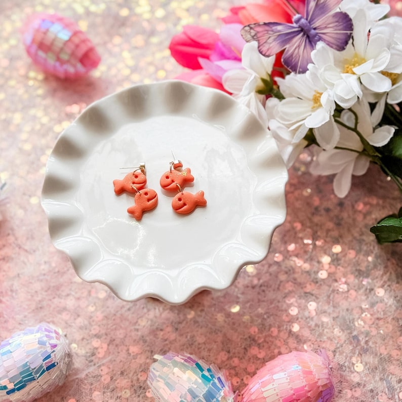 A pair of handmade goldfish cracker-inspired dangle earrings displayed on a white scalloped dish. Each earring consists of two small orange fish-shaped charms hanging vertically from gold-toned posts. The background includes pink sequins, decorative pastel eggs, artificial flowers, and a purple butterfly.