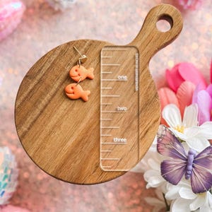 A single handmade goldfish cracker-inspired dangle earring displayed on a small wooden cutting board. The earring features two orange fish-shaped charms hanging from a gold-toned post. A clear acrylic ruler is placed next to it for size reference, measuring approximately 1.5 inches. The background includes pink sequins, artificial flowers, and a purple butterfly decoration.