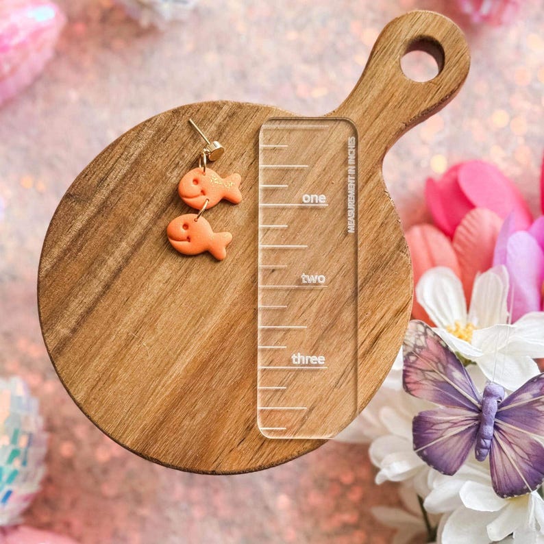 A single handmade goldfish cracker-inspired dangle earring displayed on a small wooden cutting board. The earring features two orange fish-shaped charms hanging from a gold-toned post. A clear acrylic ruler is placed next to it for size reference, measuring approximately 1.5 inches. The background includes pink sequins, artificial flowers, and a purple butterfly decoration.