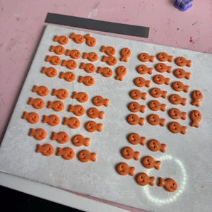 A work-in-progress display of handmade polymer clay goldfish cracker-inspired charms arranged in neat rows on a baking tile. The small orange fish-shaped pieces have smiling faces and feature a subtle shimmer. Surrounding the workspace are various clay cutters, sculpting tools, and pastel-colored craft supplies on a pink tabletop.