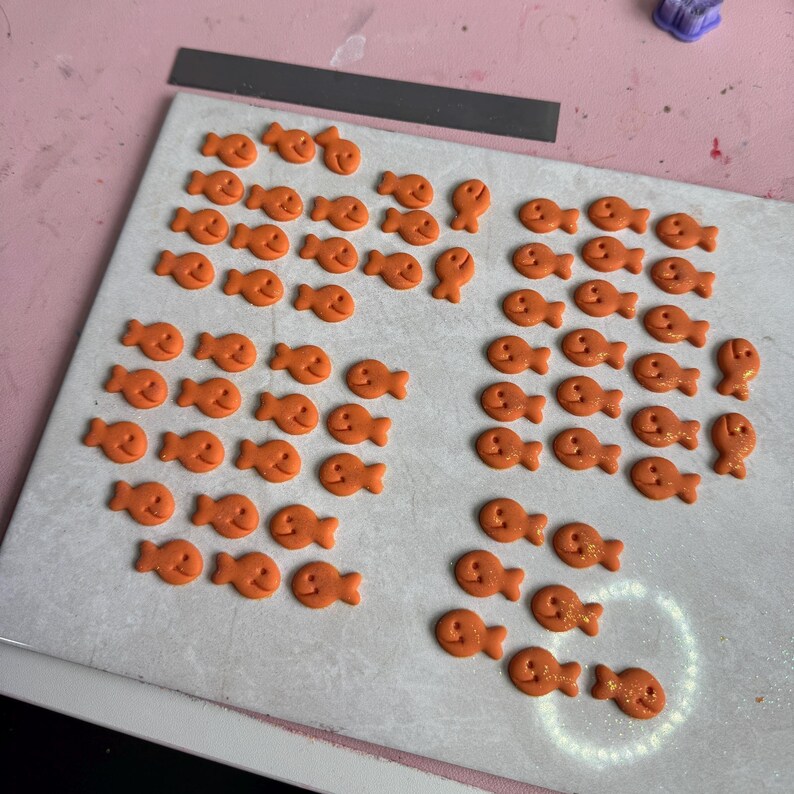 A work-in-progress display of handmade polymer clay goldfish cracker-inspired charms arranged in neat rows on a baking tile. The small orange fish-shaped pieces have smiling faces and feature a subtle shimmer. Surrounding the workspace are various clay cutters, sculpting tools, and pastel-colored craft supplies on a pink tabletop.