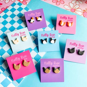 May include: A collection of cat-shaped stud earrings on colorful display cards. The earrings feature various color combinations, including black and white, orange and white, and brown and tan. Each card displays the "Nelly Rose jewelry" logo.