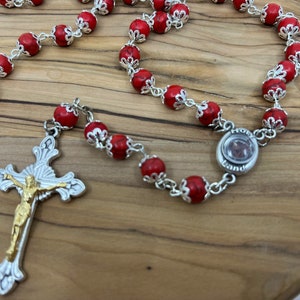 Könnte beinhalten: Eine silberne und rote Rosenkranz mit einem silbernen Kruzifix und einem runden silbernen Anhänger mit den Worten "Heiliges Land".