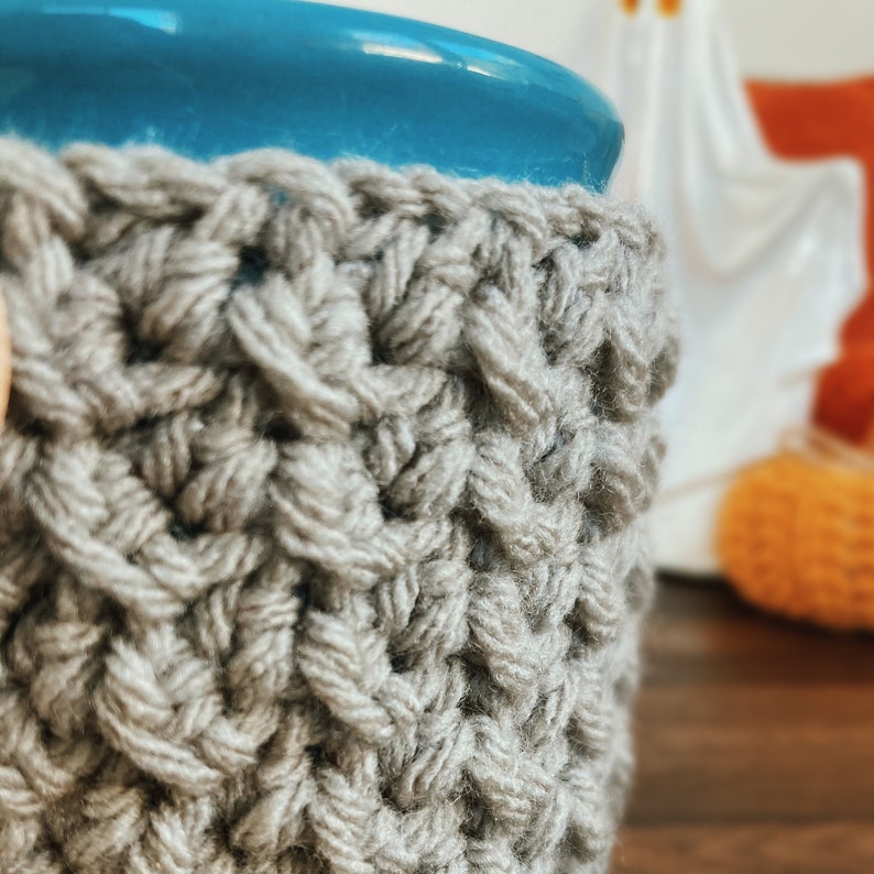 K&ouml;nnte beinhalten: Nahaufnahme einer grauen, handgestrickten Kaffeebecherh&uuml;lle. Die H&uuml;lle ist um einen blauen Keramikbecher gewickelt. Die Strickstruktur ist sichtbar, mit einem sich wiederholenden Muster. Der Hintergrund ist verschwommen, mit Andeutungen von Herbstdekorationen.
