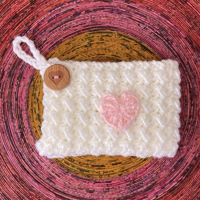 K&ouml;nnte beinhalten: Cremefarbene geh&auml;kelte Tasche mit einem rosa Herz-Applikation und einem Holzknopfverschluss. Die Tasche hat eine Schlaufe zum Aufh&auml;ngen. Das H&auml;kelmuster ist ein strukturiertes Gewebe. Ein bezaubernder handgefertigter Artikel.