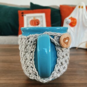 K&ouml;nnte beinhalten: Eine t&uuml;rkisfarbene Keramiktasse mit einer grauen H&auml;kelmanschette, die mit einem Holzknopf mit Herzdesign befestigt ist. Die Tasse steht auf einer Holzoberfl&auml;che, mit einem verschwommenen Hintergrund mit Halloween-Dekorationen.