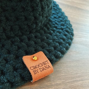 May include: Close-up of a dark green crocheted hat with a textured pattern. A small, brown leather tag with the text "CROCHET BY DANA" is attached with a gold-colored stud. The hat is handmade.