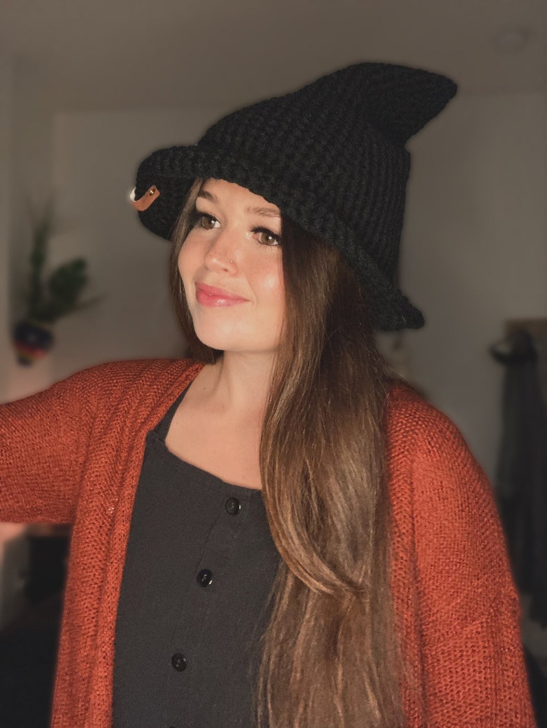 May include: A black crochet witch hat with a pointed crown and a wide brim. The hat has a small leather tag attached to the side. The hat is part of a costume or fashion accessory.