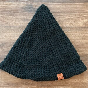 May include: A dark green, crocheted hat with a triangular shape. The hat has a small, brown leather tag with the text "CROCHET BY DANA". The hat is made of a thick yarn and has a textured appearance.