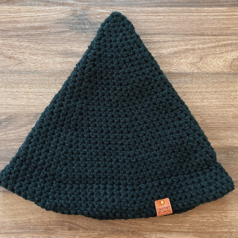 May include: A dark green, crocheted hat with a triangular shape. The hat has a small, brown leather tag with the text "CROCHET BY DANA". The hat is made of a thick yarn and has a textured appearance.