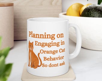 Könnte beinhalten: Weiße Keramik-Tasse mit dem Text "Planning on Engaging in Orange Cat Behavior So dont ask" in Orange. Auf der linken Seite der Tasse befindet sich eine Illustration einer orangefarbenen Katze. Die Tasse steht auf einem beigen Untersetzer.