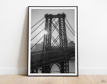 May include: Black and white photograph of a bridge in New York City. The bridge is a suspension bridge with two towers and a roadway. The sun is shining through the bridge's structure, creating a starburst effect.