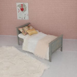 Puede incluir: Una escena de dormitorio en miniatura con una cama gris, ropa de cama blanca, una manta color melocotón y almohadas decorativas. Una alfombra de piel sintética blanca está en el suelo. Una ilustración enmarcada de una mujer con auriculares cuelga de la pared rosa.