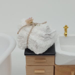Könnte beinhalten: Miniatur-Badezimmer-Set mit einer weißen Badewanne, einem Holzschrank mit einem weißen Waschbecken und goldenen Wasserhähnen sowie einem Stapel weißer Handtücher, die mit Bindfaden zusammengebunden sind. Das Set steht auf einem weißen, flauschigen Teppich.