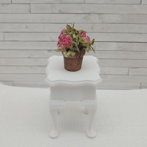 May include: A miniature white table with curved legs supports a small brown pot filled with pink flowers and green leaves. The arrangement sits on a white textured surface, against a white wooden plank backdrop.