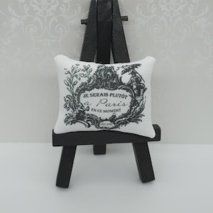 May include: A miniature white cushion with black text and a floral design, presented on a small black easel. The text on the cushion reads "Je serais plutôt Paris en ce moment."