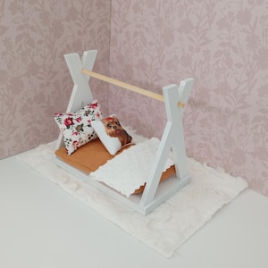 Puede incluir: Una pequeña cama blanca en forma de tipi con una barra de madera, que sostiene una cama en miniatura. Incluye un colchón marrón, una manta blanca y dos almohadas decorativas. Una almohada tiene un estampado floral y la otra presenta la cara de un perro. La cama descansa sobre una alfombra blanca y esponjosa.