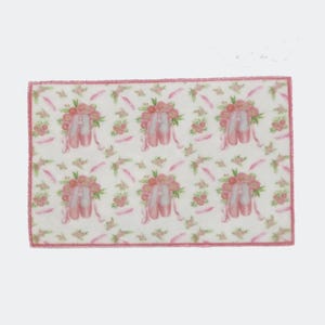 Puede incluir: Un paño rectangular blanco con un borde rosa presenta un patrón repetido de zapatillas de ballet rosas adornadas con flores. El diseño incluye plumas rosas y detalles florales, creando una estética delicada y femenina.