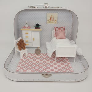 May include: Miniature bedroom scene inside a white suitcase. The scene includes a white bed with a pink pillow, a white dresser with yellow drawer pulls, a small chair with a teddy bear, and a pink and white floral rug. A framed picture hangs on the wall.