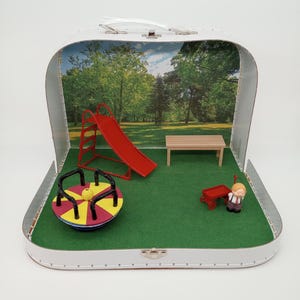 May include: A miniature playground scene inside a white suitcase. The scene includes a red slide, a wooden bench, a colorful merry-go-round, and a small red wagon. A miniature figure is also present. The background depicts a park with trees.