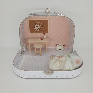 May include: A miniature dollhouse scene inside a white suitcase. The interior features a small desk, stool, corkboard with a flower design, and a floral pillow. The walls are pink with a subtle pattern.