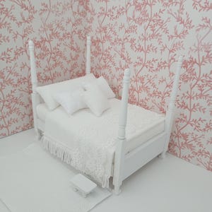 May include: Miniature white four-poster bed with bedding, pillows, and a fringed blanket. A small white rug and a side table are in front of the bed. The background features a pink and white floral wallpaper.