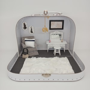 May include: A miniature dollhouse scene set within a white suitcase. The interior includes a black and white patterned floor, a fluffy white rug, a striped armchair, a vanity with a mirror, and framed artwork. A small chandelier hangs from the top.