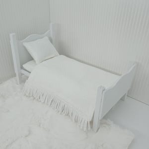 May include: A miniature white bed with a matching pillow and blanket. The blanket has a fringe along the bottom edge. The bed sits on a white, fluffy rug. The bed frame is made of wood.