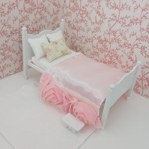 May include: Miniature white bed with a pink satin comforter and a decorative pink rose-like trim. Two white pillows and a floral pillow rest on the bed. A white rug and a small white nightstand are in front of the bed. The background is a pink floral wallpaper.