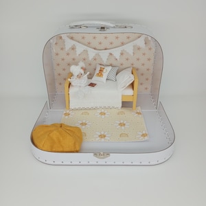 May include: A white suitcase opens to reveal a miniature bedroom scene. Inside, a small bed with pillows and a teddy bear pillow sits on a daisy-patterned rug. A yellow hat rests in the suitcase, and a banner hangs above the bed.