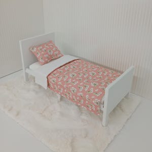 May include: A miniature white bed with a pink floral patterned comforter and pillow. The bed sits on a fluffy white rug. The bedding features a delicate white flower design on a pink background, creating a soft, inviting aesthetic.