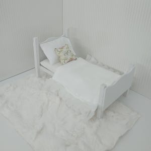 Könnte beinhalten: Ein kleines weißes Bett mit weißer Bettwäsche und einem kissen mit floralem Muster. Unter dem Bett liegt ein flauschiger weißer Teppich. Das Bett befindet sich in einem weißen Raum. Das Bett ist etwa 30 cm lang.