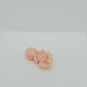 以下が含まれることがあります： 仰向けに寝ている赤ちゃんの小さな薄ピンク色のフィギュア。腕と脚は丸まっています。赤ちゃんの頭は横を向き、目は閉じられています。フィギュアは白い表面にあります。