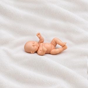 May include: A small, light-skinned baby doll in a reclining position with arms and legs slightly bent. The doll features molded facial details, including closed eyes, a small nose, and a mouth, with a smooth, matte finish throughout its body.