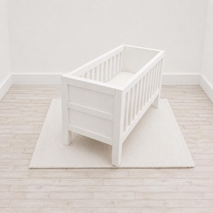 Op de afbeelding: Een wit houten wiegje met een rechthoekig frame en verticale spijlen aan alle vier de zijden. Het wiegje heeft een stevige bodem en vier robuuste poten, met een effen wit matras zichtbaar in de binnenkant van de constructie.