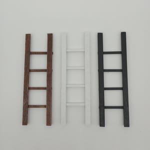 Op de afbeelding: Drie miniatuur decoratieve ladders in verschillende kleuren: bruin, wit en zwart. Elke ladder heeft drie sporten en een eenvoudig ontwerp. De ladders zijn waarschijnlijk voor woondecoratie of knutselprojecten.